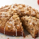 Apple Cider Coffee Cake with crumb topping and light cider glaze, one slice cut, moist and tender fall dessert on a white surface.