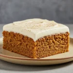 A square slice of spice cake made from a cozy chai cake recipe sits on a beige plate, topped with creamy frosting and a dusting of chai spice for fall baking.
