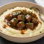 A shallow beige bowl of mashed potatoes topped with glossy Beef Tips in rich brown gravy, showing Instant Pot Beef Tips with Gravy styled like an Instant Pot stew and perfect for fans of Beef Tips And Gravy and simple Beef Tip Recipes.