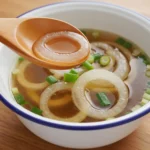 A white bowl filled with clear amber Japanese Onion Soup shows round onion slices and chopped scallions, with a wooden spoon lifting an onion ring, highlighting a light Japanese Soup styled as a Clear Soup made from a delicate Japanese Broth Soup base.