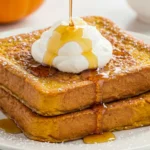 A close-up of two thick slices of Fall Pumpkin French Toast on a white plate, golden French Toast with maple syrup, a swirl of whipped pumpkin cream, cinnamon dust, and pumpkins in the background for a cozy fall breakfast.
