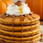 A tall stack of Pumpkin Spice Pancakes sits on a white plate, drenched in maple syrup and topped with whipped cream and pecans, a cozy Pancakes moment that looks like Fluffy Pumpkin Pancakes from a Pumpkin Pancake Recipe for the perfect Fall Breakfast and a keeper for Fall Recipes.