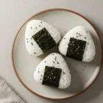 Close-up of Japanese Tuna Onigiri, triangle sushi rice ball with tuna mayo filling, black sesame seeds, and nori wrap held in hand.