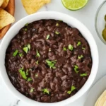 Bowl of refried black beans topped with cilantro, surrounded by tortilla chips, lime, jalapeños, and tortillas, easy homemade Mexican side