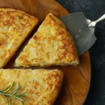 Authentic Tortilla de Patatas - Restaurant-Quality Results At Home 5 Tortilla de Patatas, traditional Spanish potato omelette on a wooden board with a cut slice